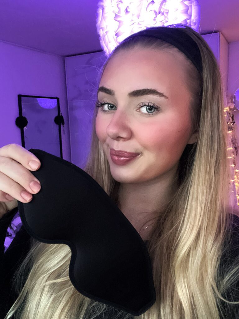 Selfie af ung kvinde med langt blondt hår og pandebånd holder sort sovemaske i lilla oplyst værelse