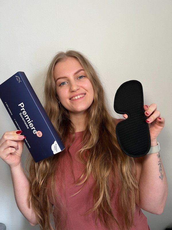 Ung kvinde smilende holder Premiere-sovemaskepakke i den ene hånd og sort sovemaske i den anden