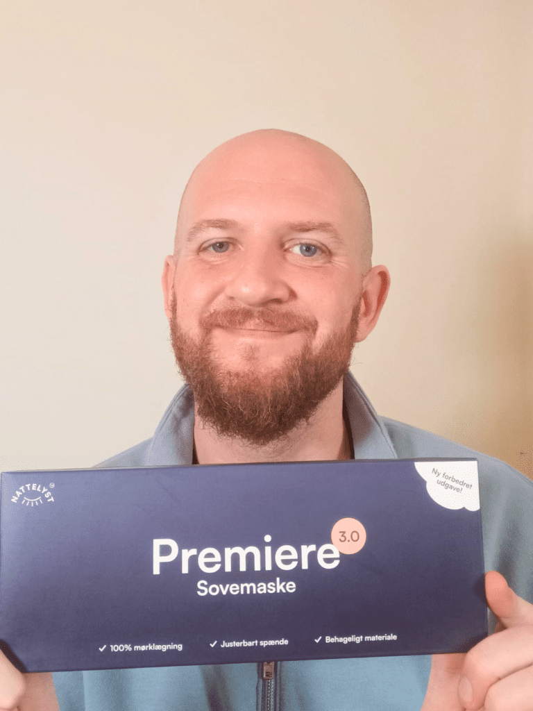 Smilende mand med skæg holder Premiere 3.0 sovemaskeæske i mørkeblå