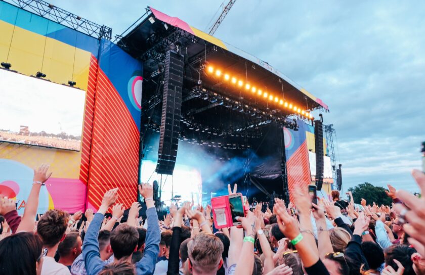 Stor udendørs musikfestivalscene under aftenhimmel, publikum med hænder i vejret