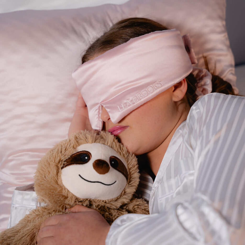 Person i lyserød sovemaske og stribet pyjamas krammer blød dovendyrbamse på rosa silkepude