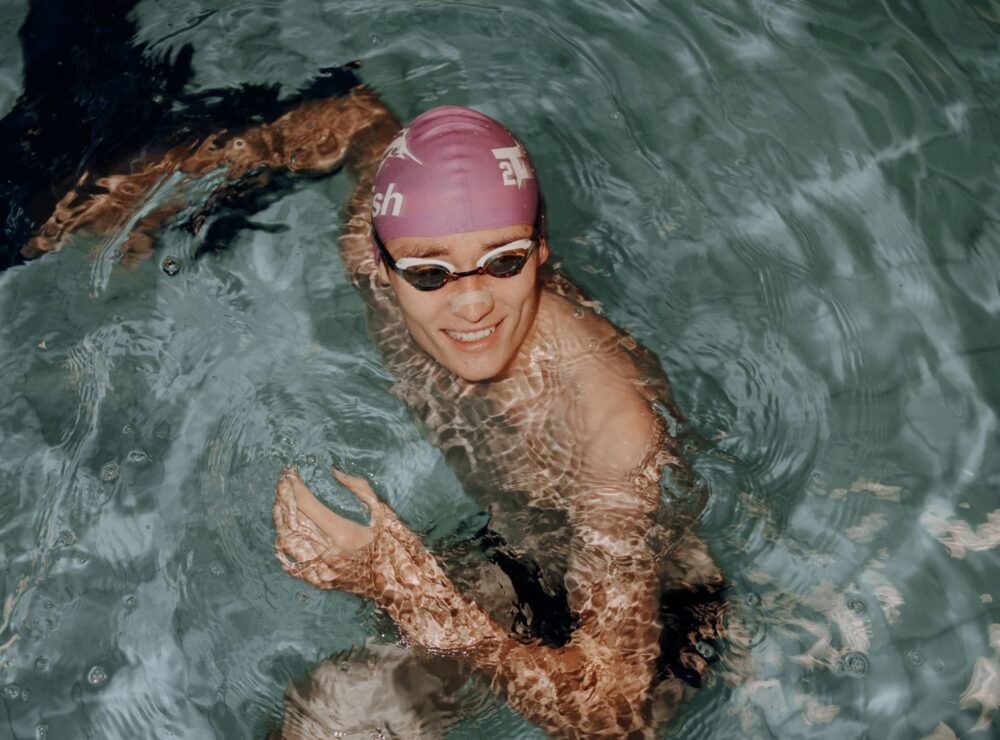 Smilende svømmer med lilla badehætte og svømmebriller i pool, vandreflekser over arm og skuldre